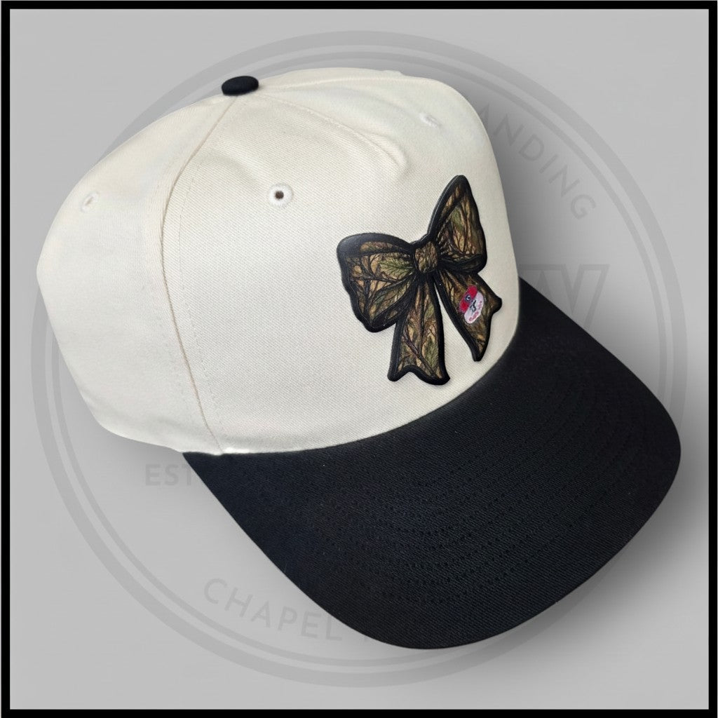 Two-tone baseball cap with cream crown, black brim, and embroidered bow patch