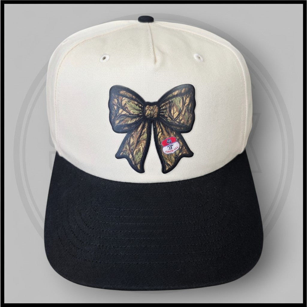 Two-tone baseball cap with cream front panels, black brim, and centered bow graphic