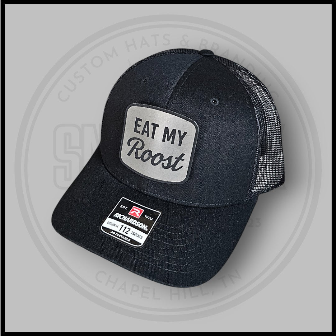 Dark charcoal trucker cap, side angle, with Eat My Roost front patch and Richardson badge