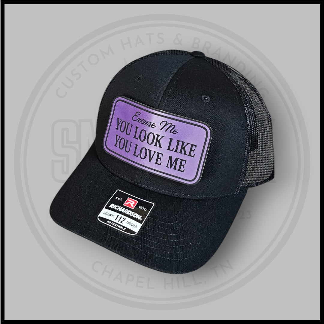 Black trucker cap with purple-bordered front patch text 'Excuse Me You Look Like You Love Me'