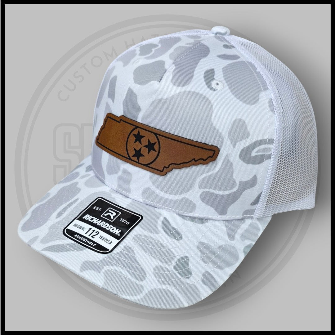 Light gray duck camo trucker cap with brown rectangular logo patch and mesh back